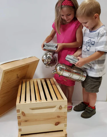 Two children playing with a wooden bee hive and metal tools on a white background